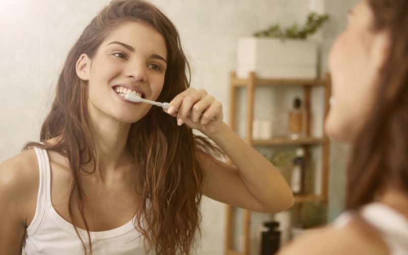 A woman avoiding teeth mistakes while brushing in front of a mirror.