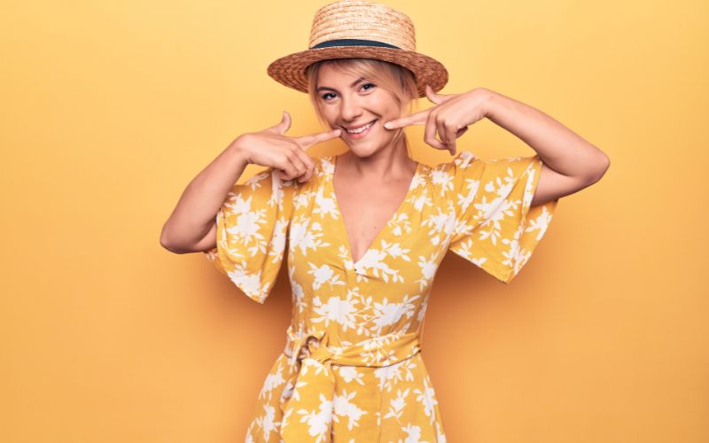 Beautiful blonde woman on vacation wearing summer hat and dress over yellow background smiling cheerful showing and pointing with fingers teeth and mouth