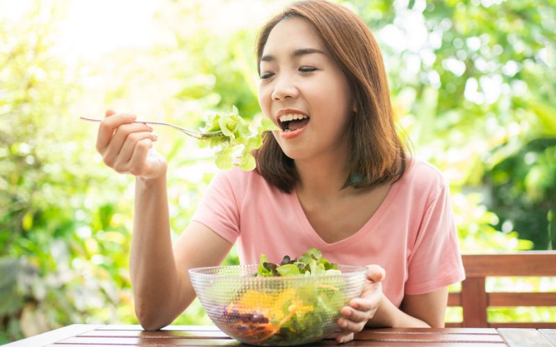 A young woman eating a bowl of salad and following 5 tips to maintain dental health.
