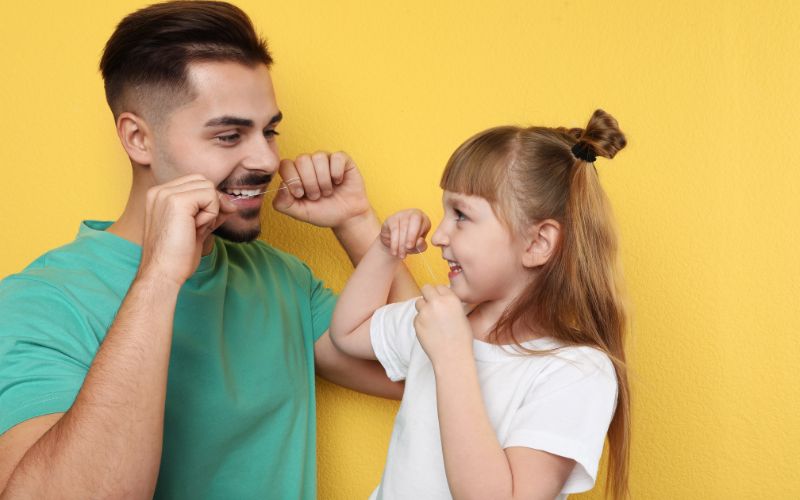 A man teaching a little girl flossing techniques against a yellow wall.