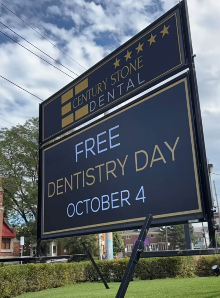 A large outdoor sign for Century Stone Dental, a trusted dentist in Hamilton, advertises “Free Dentistry Day October 4” against a backdrop of trees, sky, and nearby buildings.