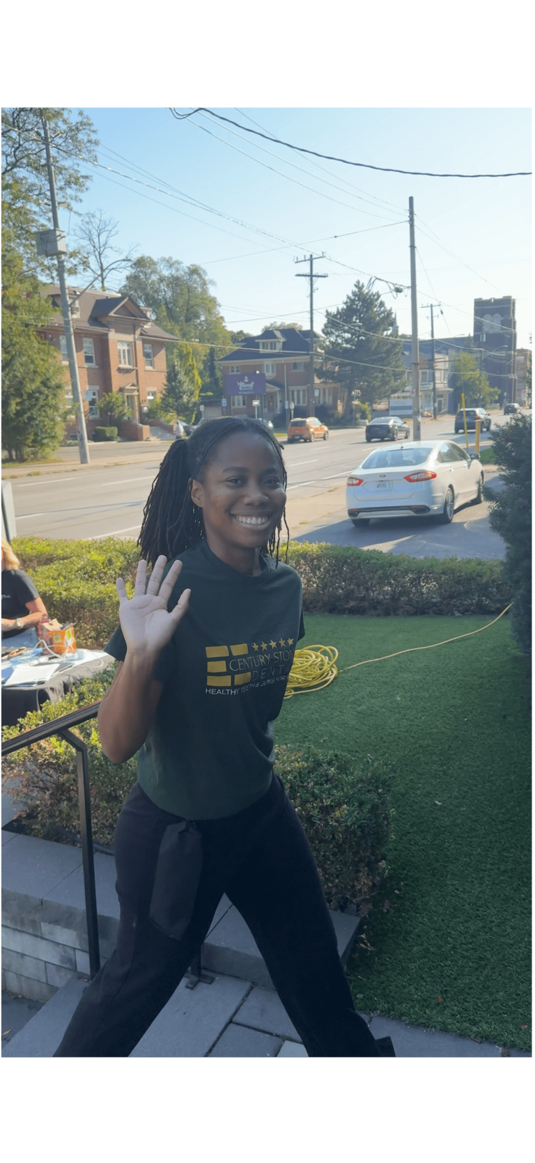 A woman stands outside on a sidewalk, smiling and waving at the camera. Cars and houses are visible in the background on a sunny day.