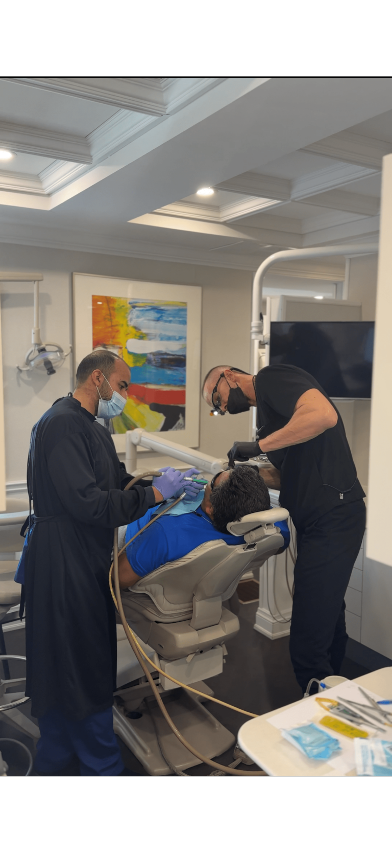 Two dental professionals wearing masks and gloves perform a procedure on a patient reclining in a dental chair inside a modern dental office.