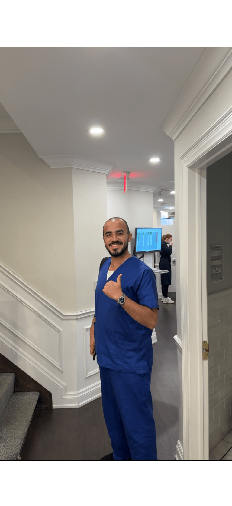 A man in blue medical scrubs stands indoors, smiling and giving a thumbs-up. A person in the background works at a desk near a wall-mounted monitor.