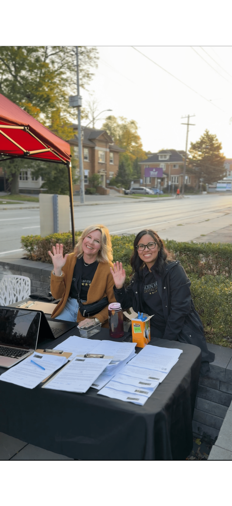 Two women sit at an outdoor table with clipboards, a laptop, and drinks, smiling and waving at the camera. Houses and a street are visible in the background.