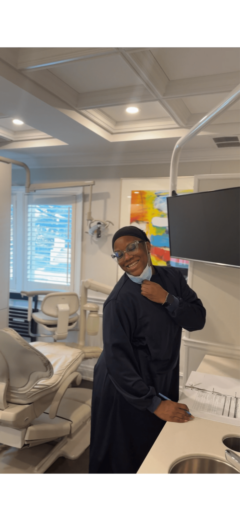 Person in dark scrubs and glasses smiles while standing next to a dental chair in a brightly lit dental office.
