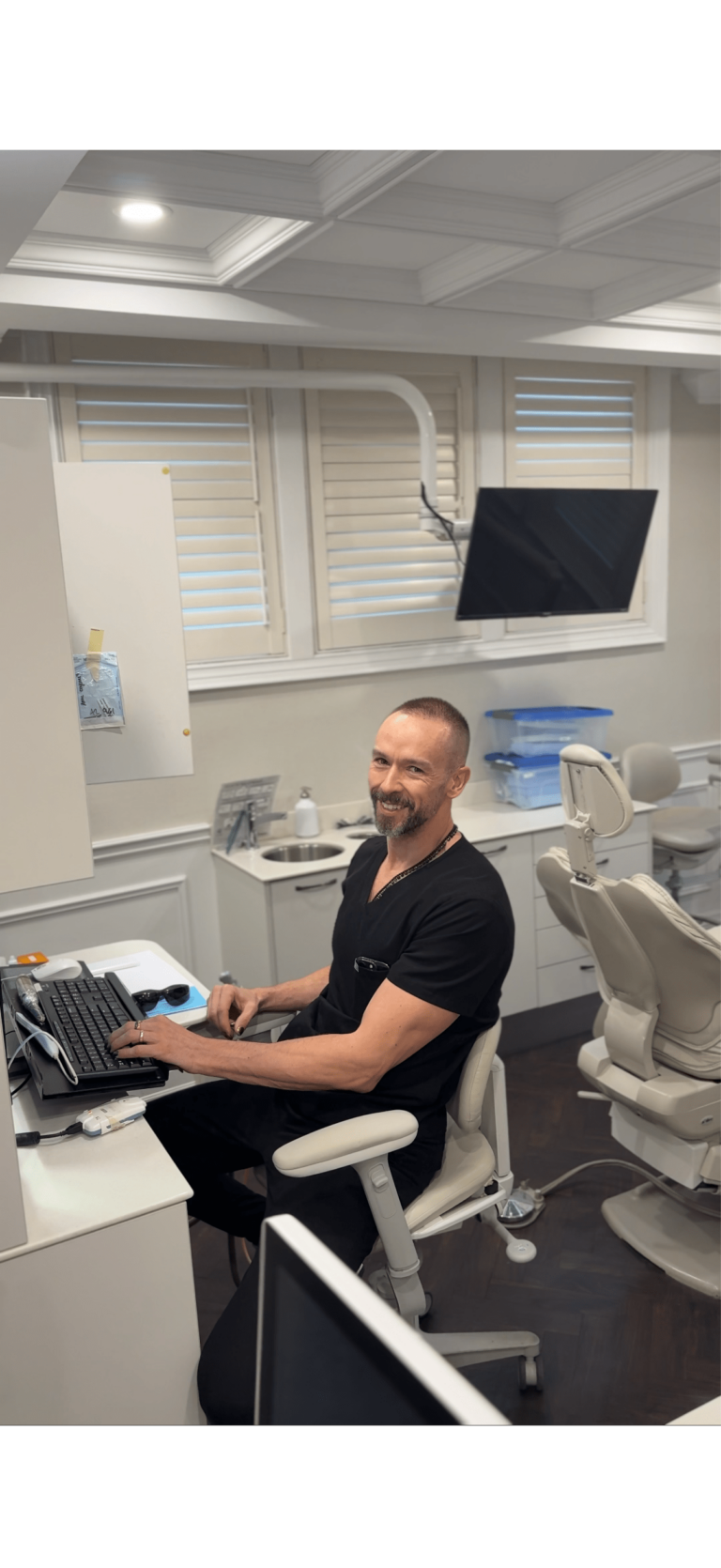 A man in black scrubs sits at a desk in a dental office, typing on a keyboard and smiling at the camera. Dental equipment and a chair are visible in the background.