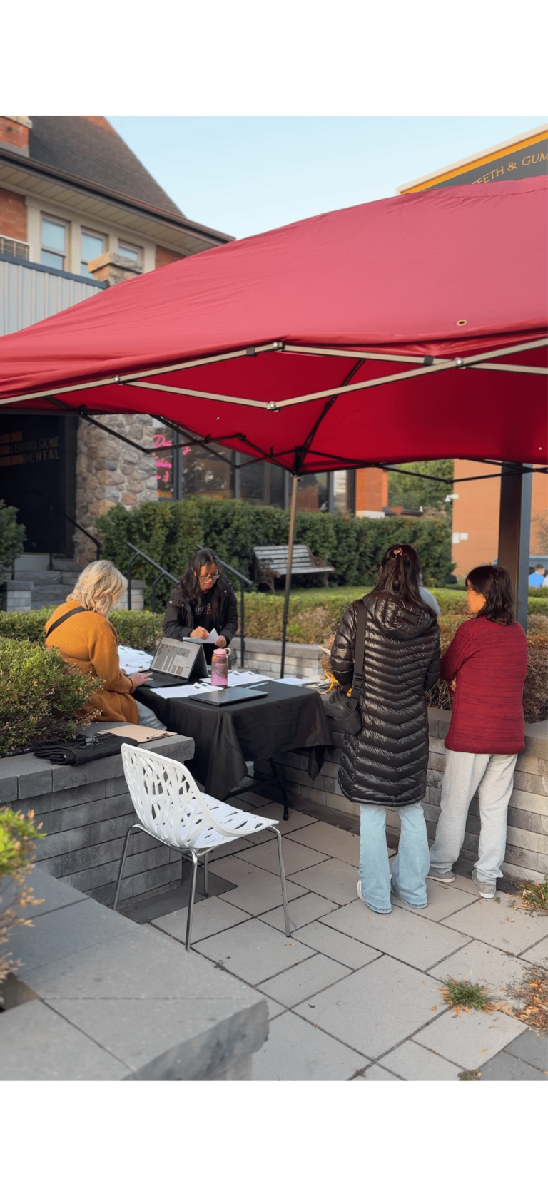 Four people interact at an outdoor table under a red canopy near a building, with paperwork and water bottles on the table.
