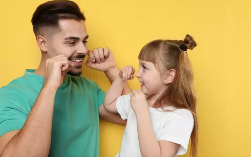 A man teaching a little girl flossing techniques against a yellow wall.