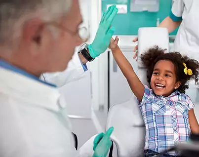 A young girl laughs while giving a high five to a children's dentist wearing gloves in a clinic, with a nurse standing in the background.