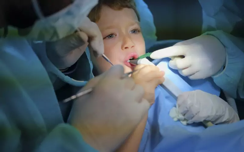 A child receives dental treatment while lying on a dental chair, being attended to by dentists wearing gloves and surgical attire.