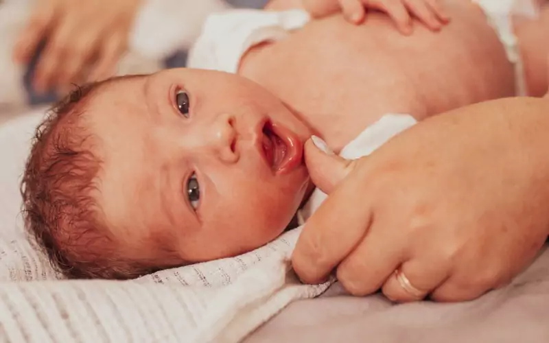 A newborn baby lies on a blanket while an adult hand gently touches its chin. The baby looks up with an open mouth and wide eyes.