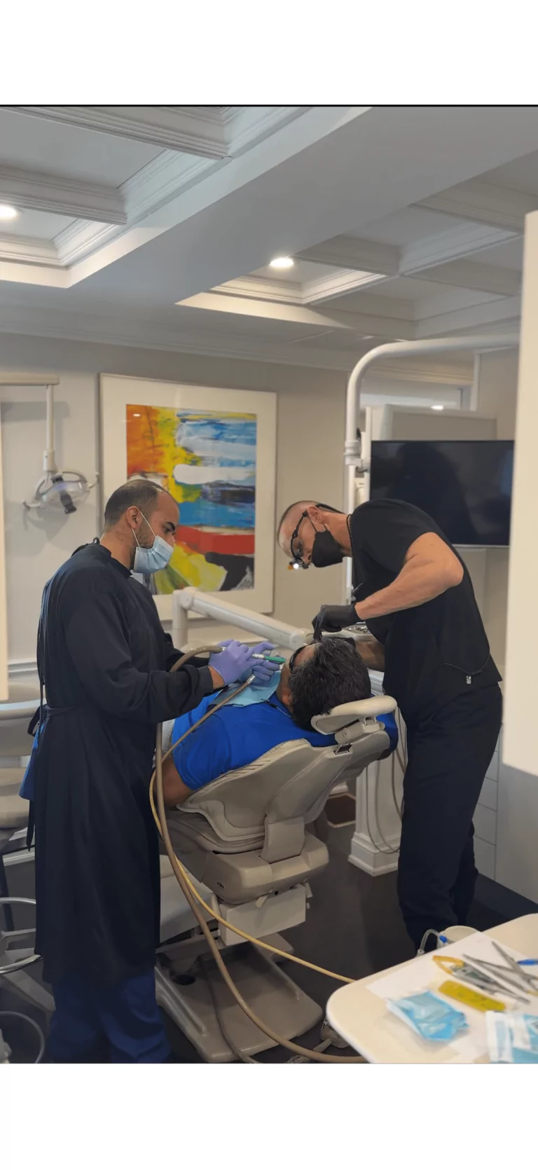 Two dental professionals wearing masks and gloves perform a procedure on a patient reclining in a dental chair inside a modern dental office.