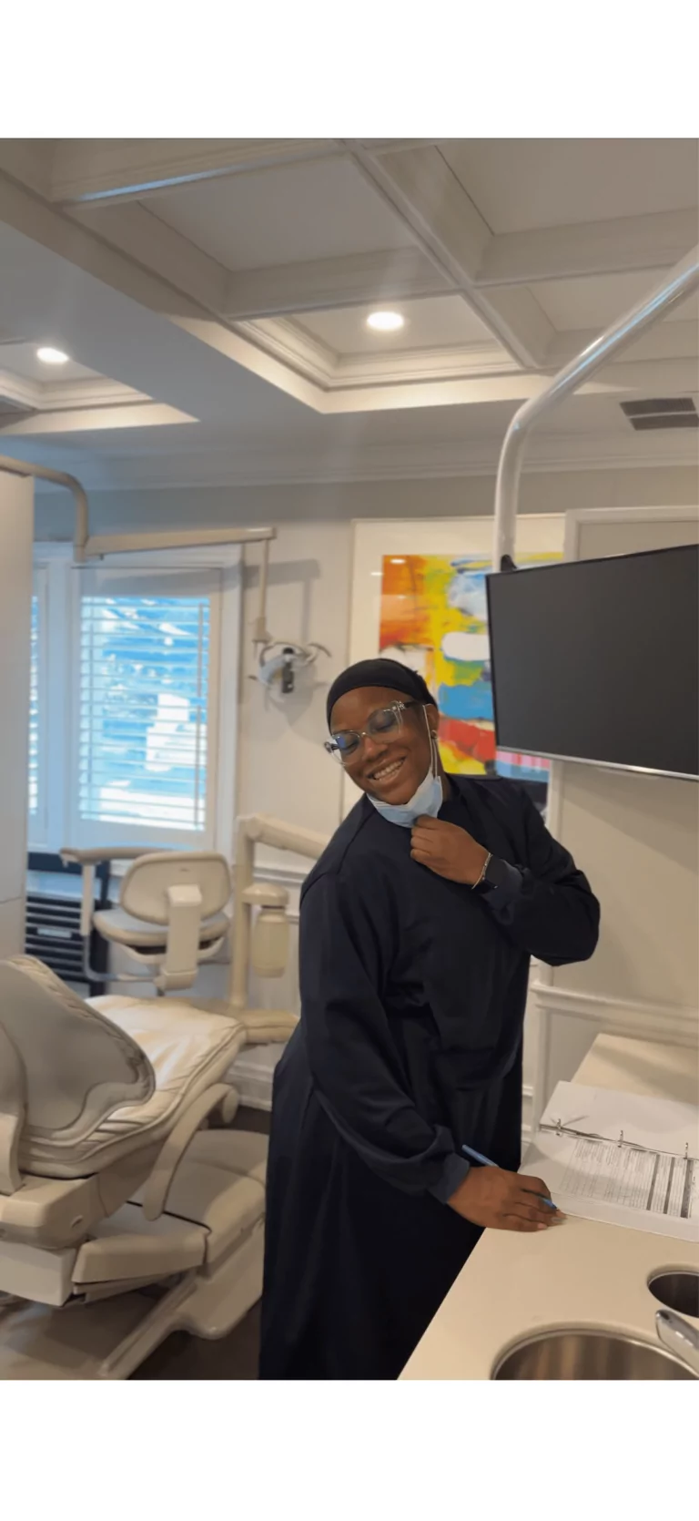Person in dark scrubs and glasses smiles while standing next to a dental chair in a brightly lit dental office.