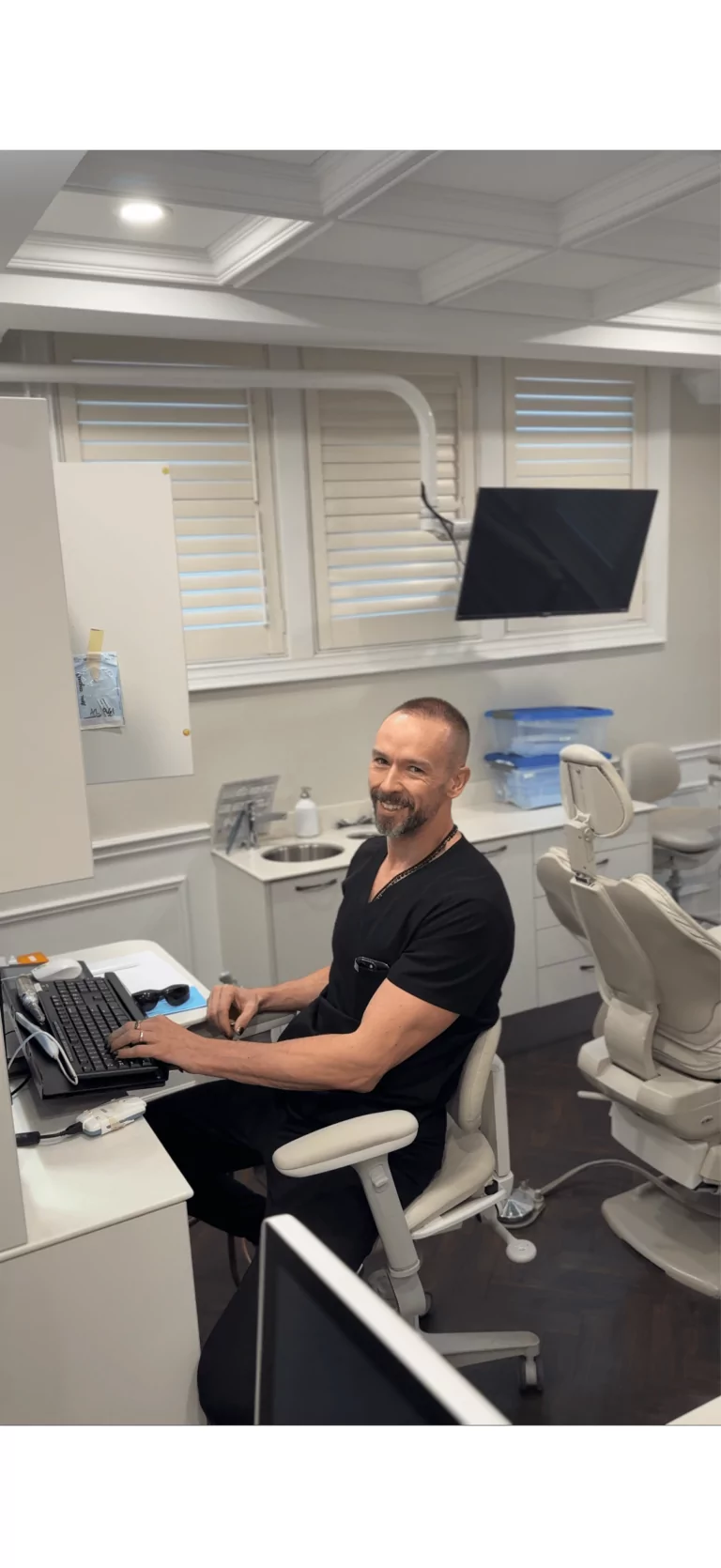 A man in black scrubs sits at a desk in a dental office, typing on a keyboard and smiling at the camera. Dental equipment and a chair are visible in the background.